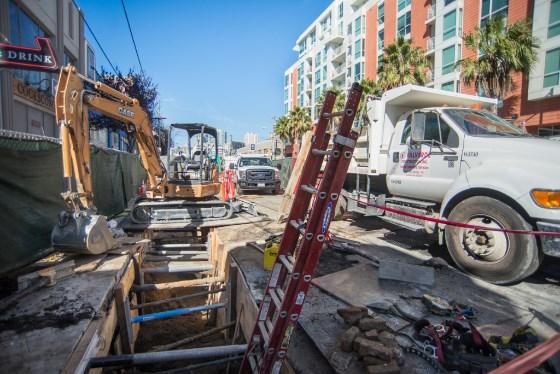 Construction equipment and vehicles on urban street with apartment buildings during Central Subway project work.