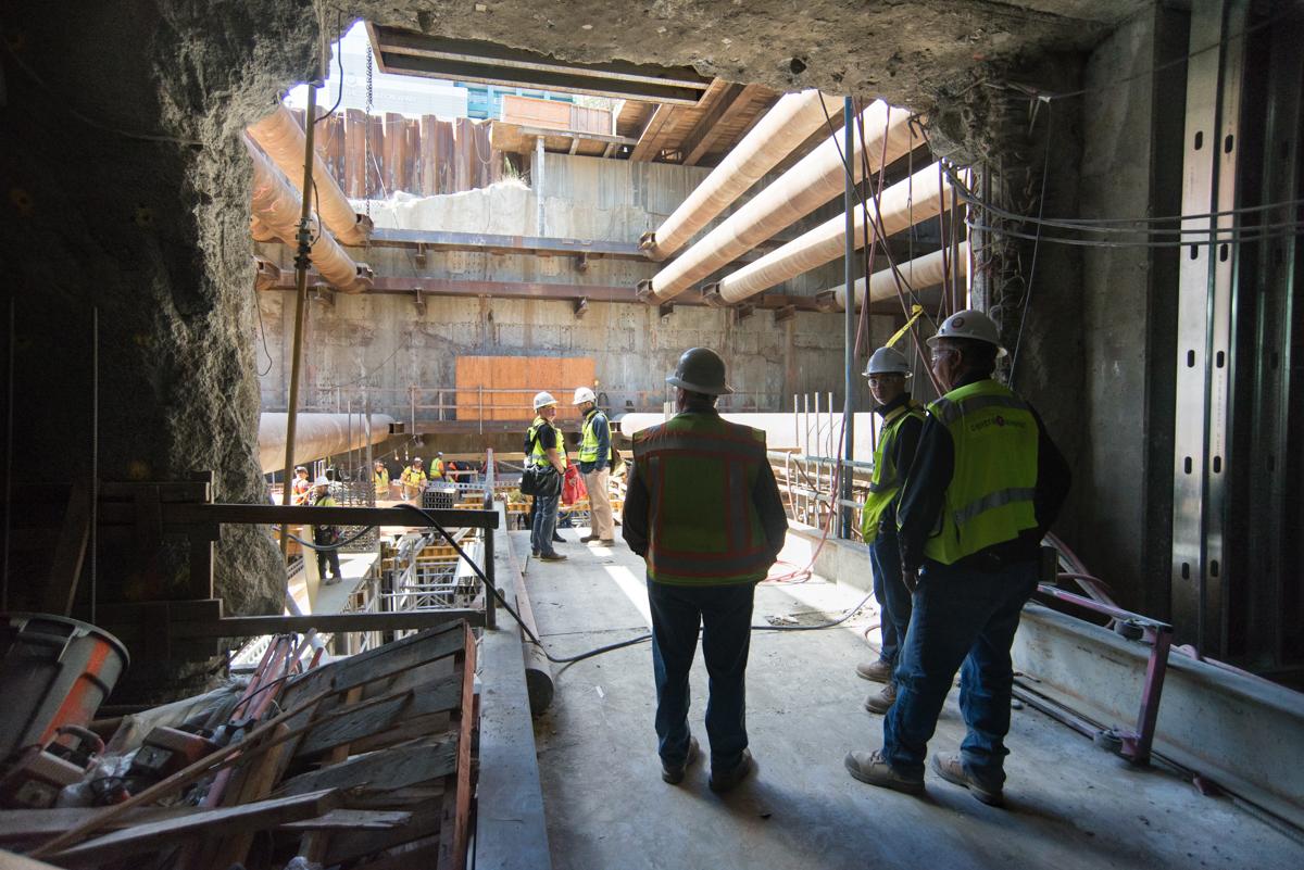 Construction workers in safety vests discuss work at an underground transit station portal with exposed concrete and wooden beams.