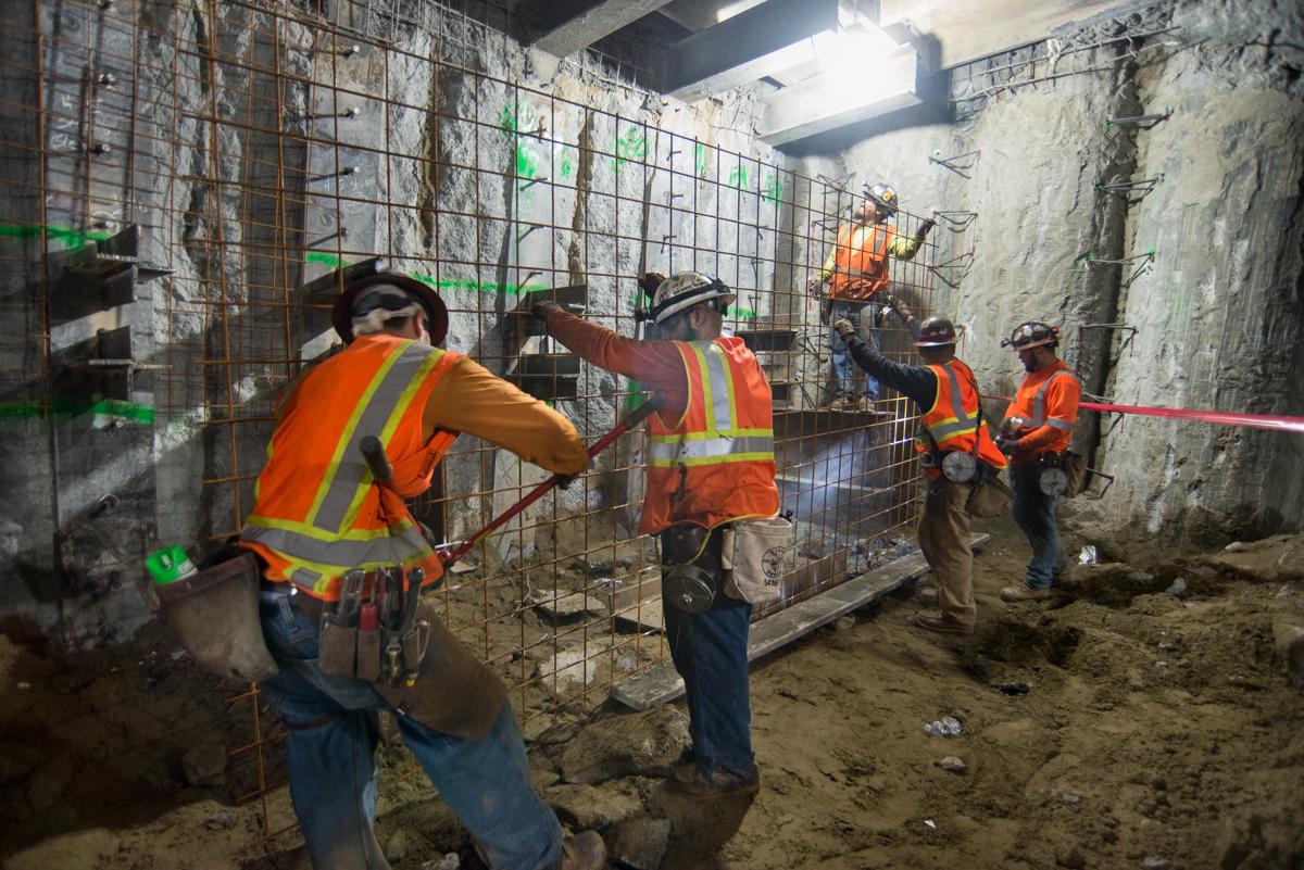 Construction workers in safety vests install steel mesh reinforcement on station box wall during transit project.