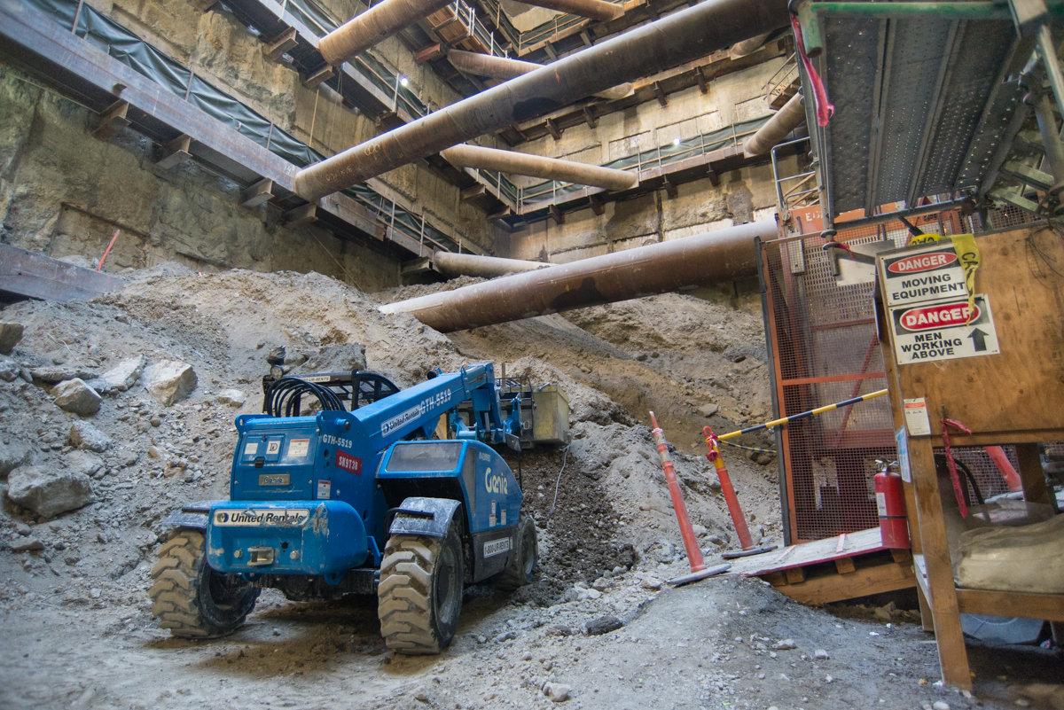 Blue excavator removing concrete in underground construction site with steel support beams overhead.