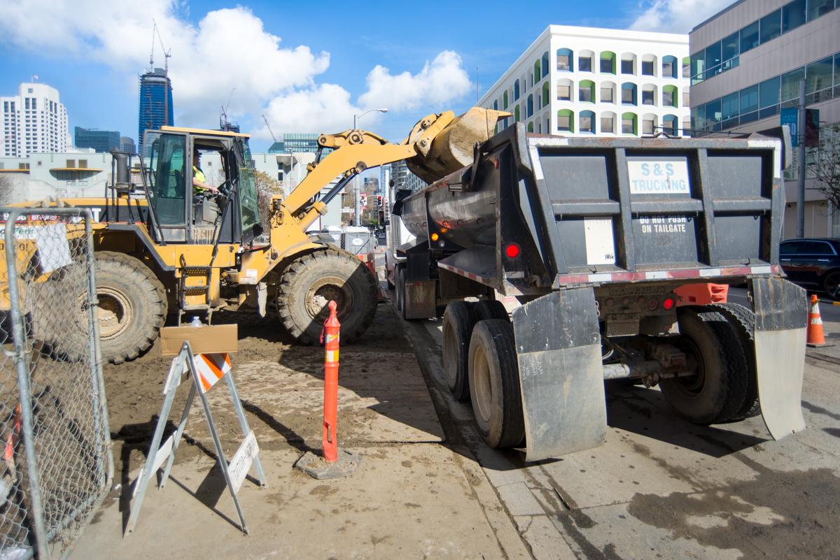 Yellow wheel loader dumps excavated material into dump truck at urban construction site.