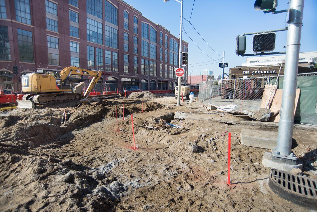 Construction excavation on 4th Street with yellow excavator, red stakes, and utility infrastructure visible.