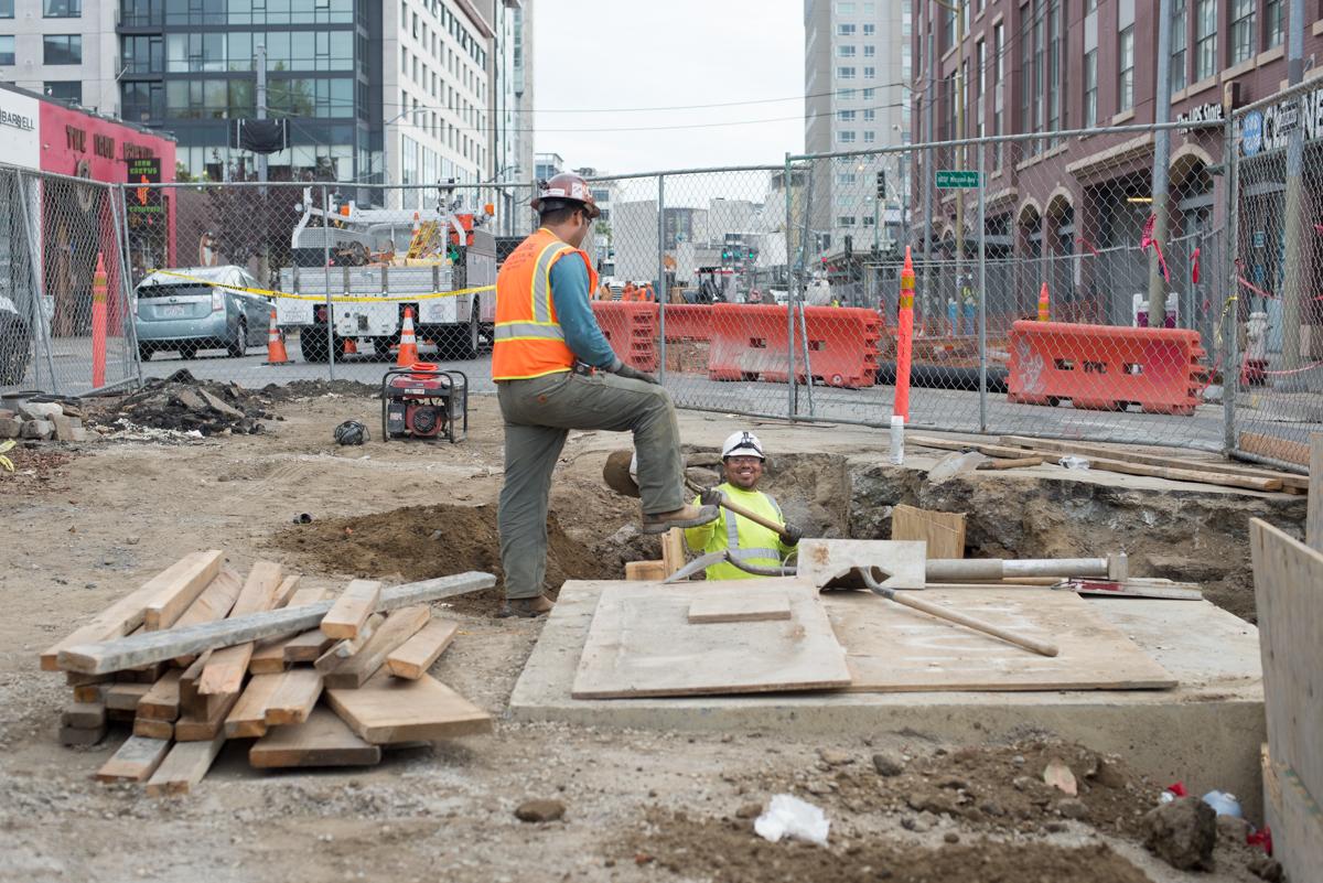 Two construction workers install utilities in a trench at an urban street intersection surrounded by fencing and equipment.