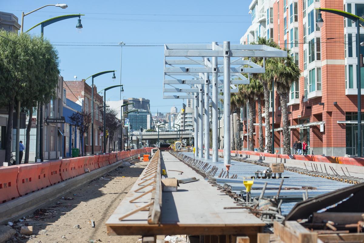 Construction of Central Subway surface station with steel framework structures and red safety barriers along urban street.