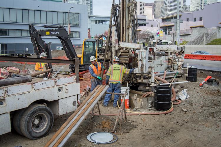 Construction workers in safety vests operate drilling equipment on an urban street surrounded by buildings and parked vehicles.