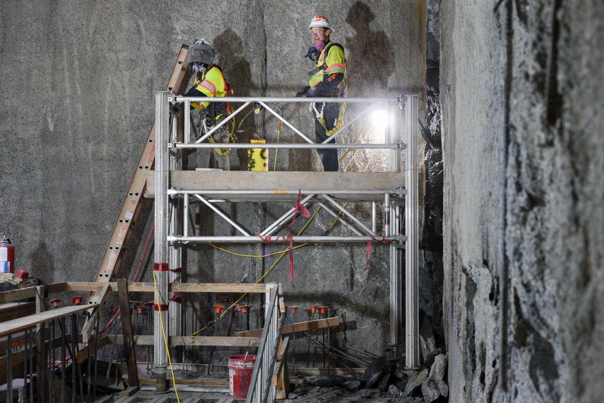 Two construction workers in safety gear on scaffolding removing concrete from interior slurry walls of a transit headhouse structure.