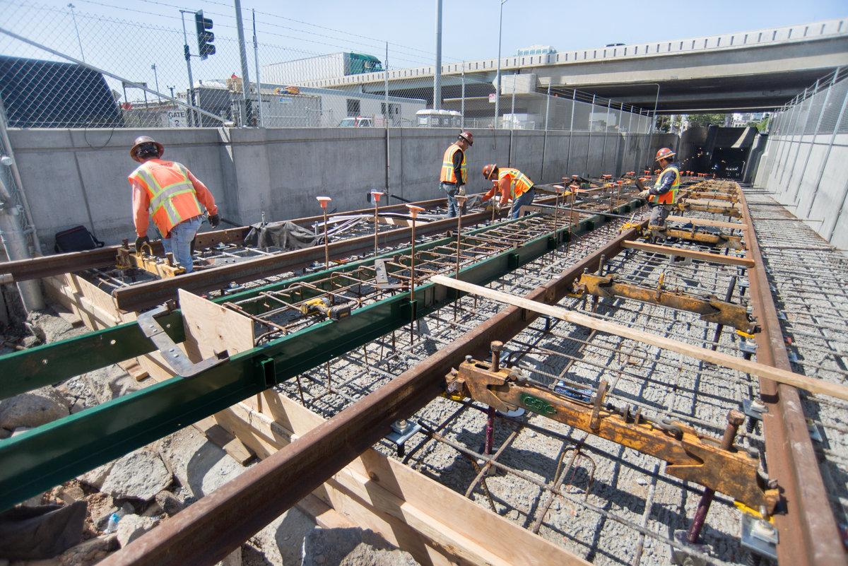 Construction workers in safety vests install railroad tracks and rebar inside a tunnel portal for the Central Subway project.