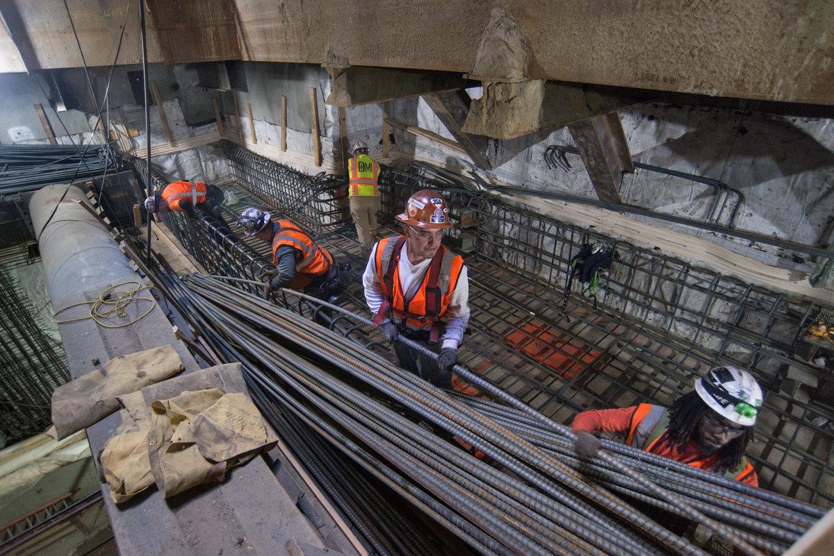 Workers in safety gear place rebar reinforcement steel at a construction site beneath an urban intersection.