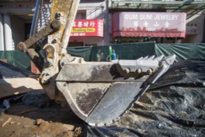 Excavator bucket positioned at construction site with street-level storefronts visible in background.