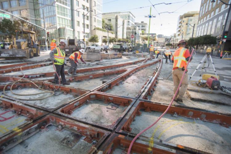 Construction workers in safety vests install streetcar rails on concrete bed in urban street.