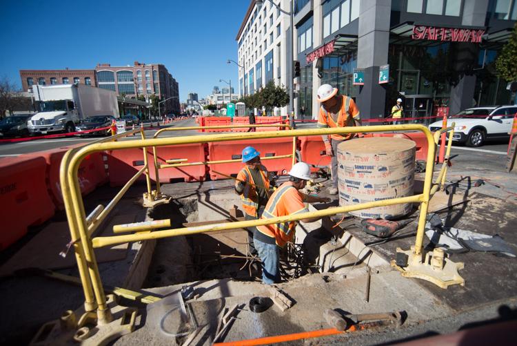 Construction workers in safety gear working in an excavated street pit with yellow safety barriers and equipment visible.
