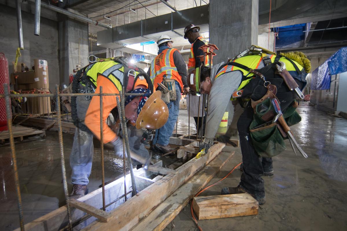Construction workers in safety vests build concrete forms inside an underground transit station platform.