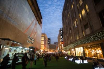 Shoppers and pedestrians walk through a downtown street decorated with holiday banners and green turf, lined with illuminated storefronts at dusk.