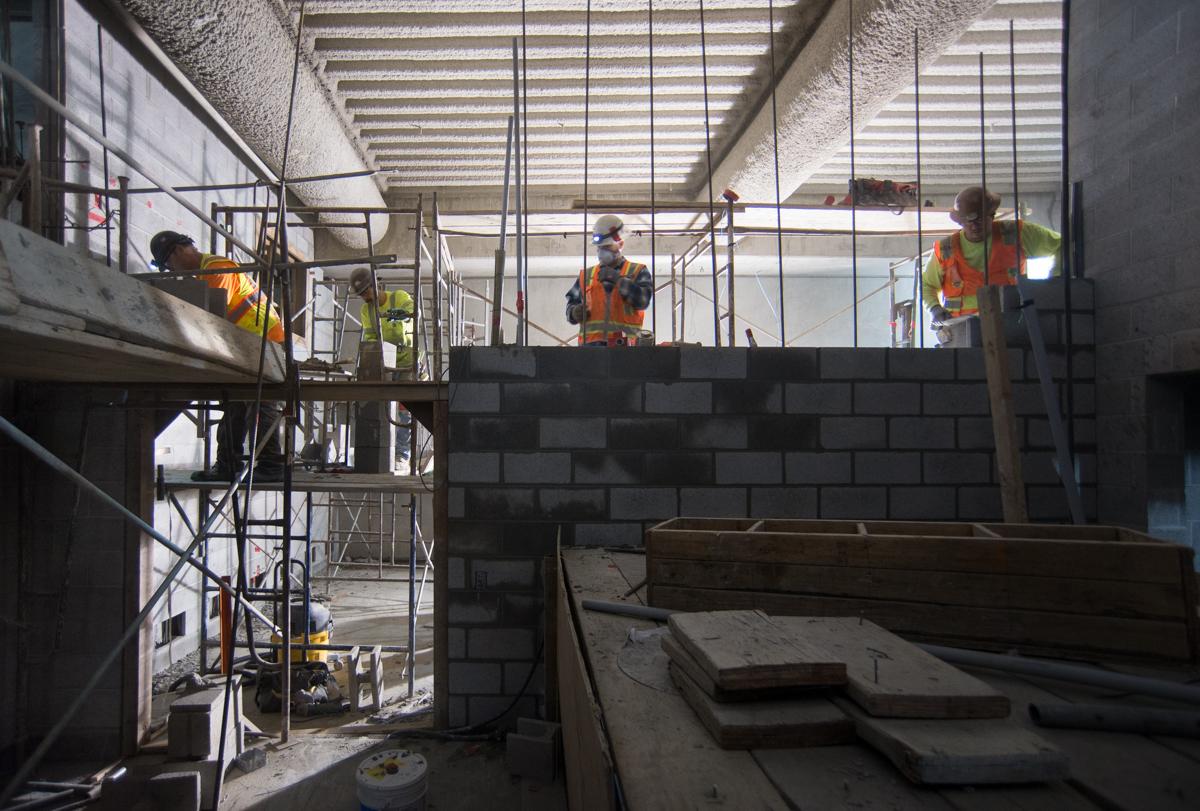 Construction workers in safety vests build a cinderblock wall on a mezzanine level with scaffolding and structural elements visible.