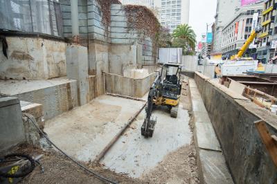 Construction equipment in excavated pit on urban street surrounded by concrete barriers and buildings.