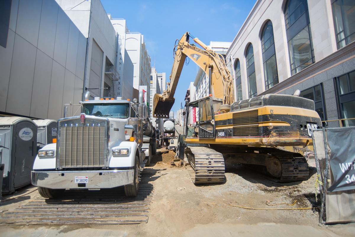 Construction site between buildings with excavator loading material onto white truck on sandy ground.