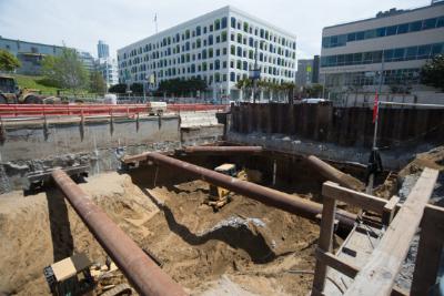Construction site with excavated trench, steel support structures, and urban buildings in background.