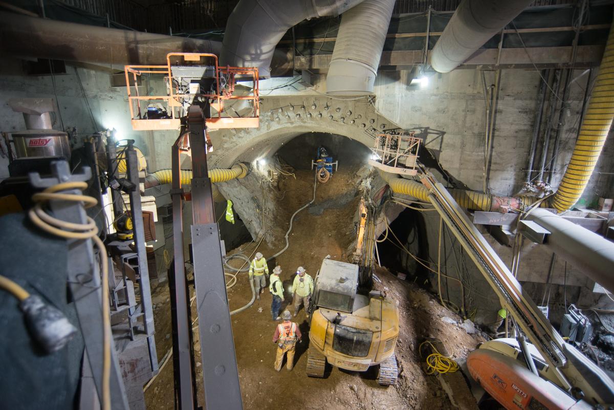 Underground transit station construction showing excavated archway entrance with workers, equipment, and support structures in concrete tunnel.