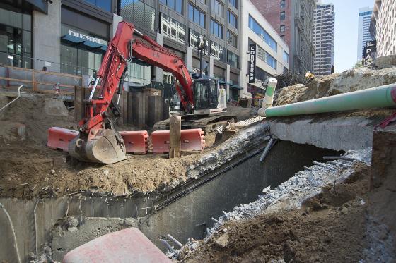 Red excavator operating in urban construction site near Powell Station with green pipe and concrete barriers visible.