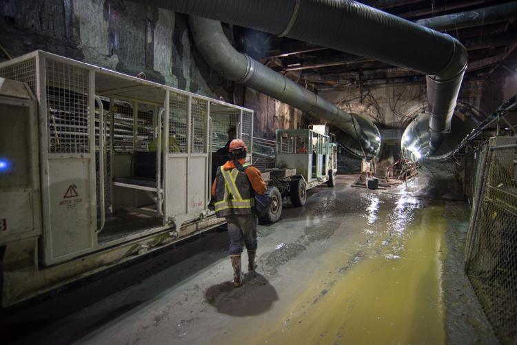Worker in hard hat and safety vest stands in wet tunnel with mining supply vehicle and large industrial pipes overhead.