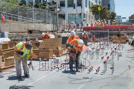 Construction workers in safety vests installing underground utility pipe supports at a transit station site.
