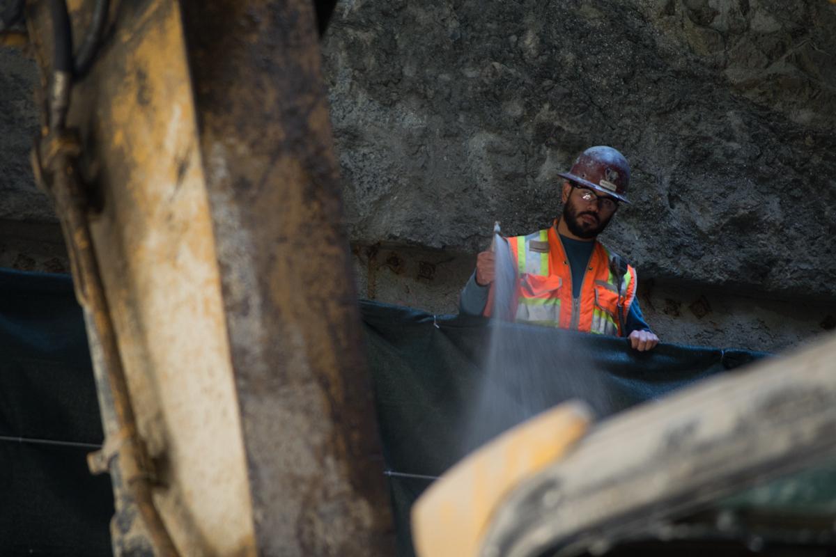 Worker in hard hat and safety vest spraying water in underground tunnel excavation.