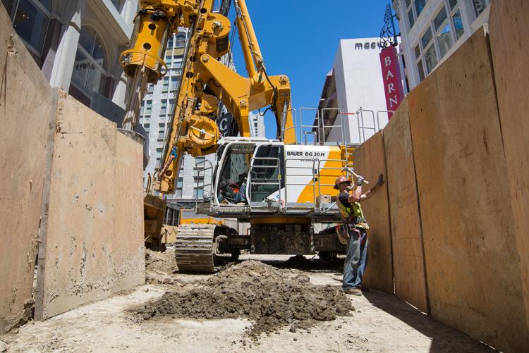 Yellow excavator with boom extended in narrow urban construction site between concrete barriers, with high-rise buildings visible above.