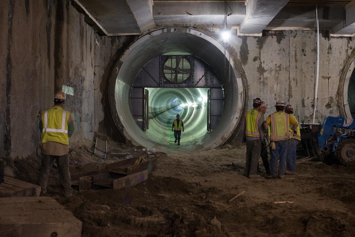 Workers in safety vests stand near a large circular tunnel portal during underground transit construction.