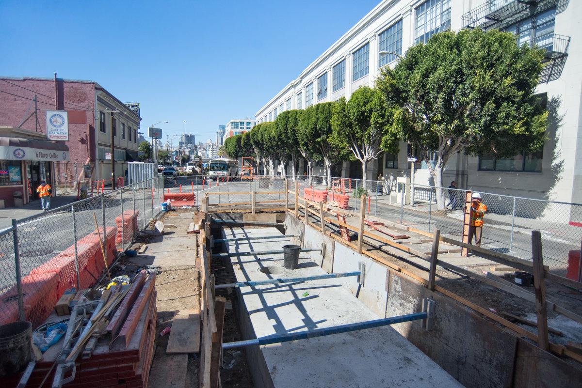 Construction site showing underground infrastructure work with concrete foundation, wooden framing, and safety fencing along urban street.