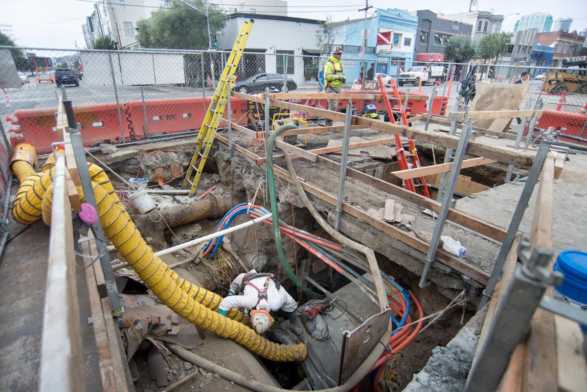 Construction workers connect sewer lines at excavation site on city street with equipment and safety barriers.