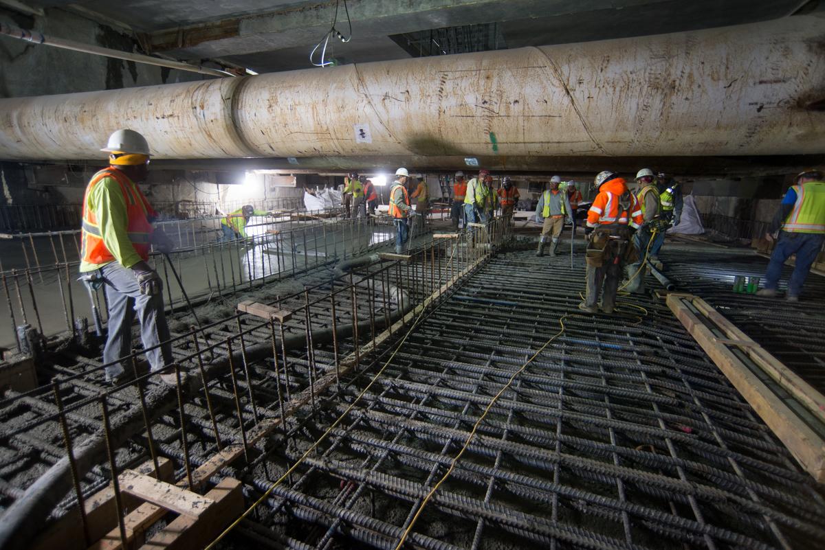 Construction workers pour concrete for transit station foundation beneath large pipe infrastructure.