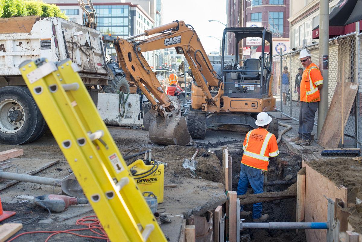 Construction crew excavating underground utilities on city street with heavy equipment and safety barriers.