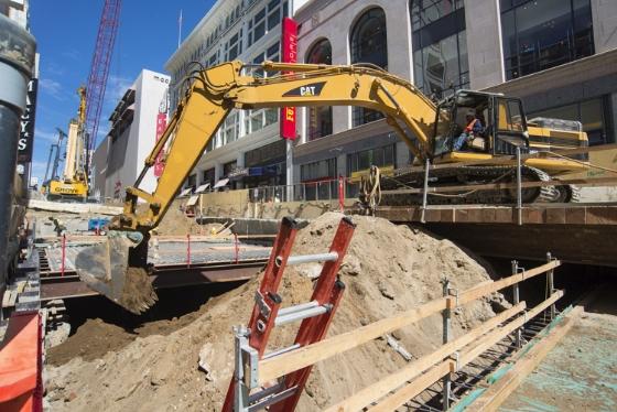 Yellow excavator on Stockton Street construction site with roof decking visible in excavation pit.