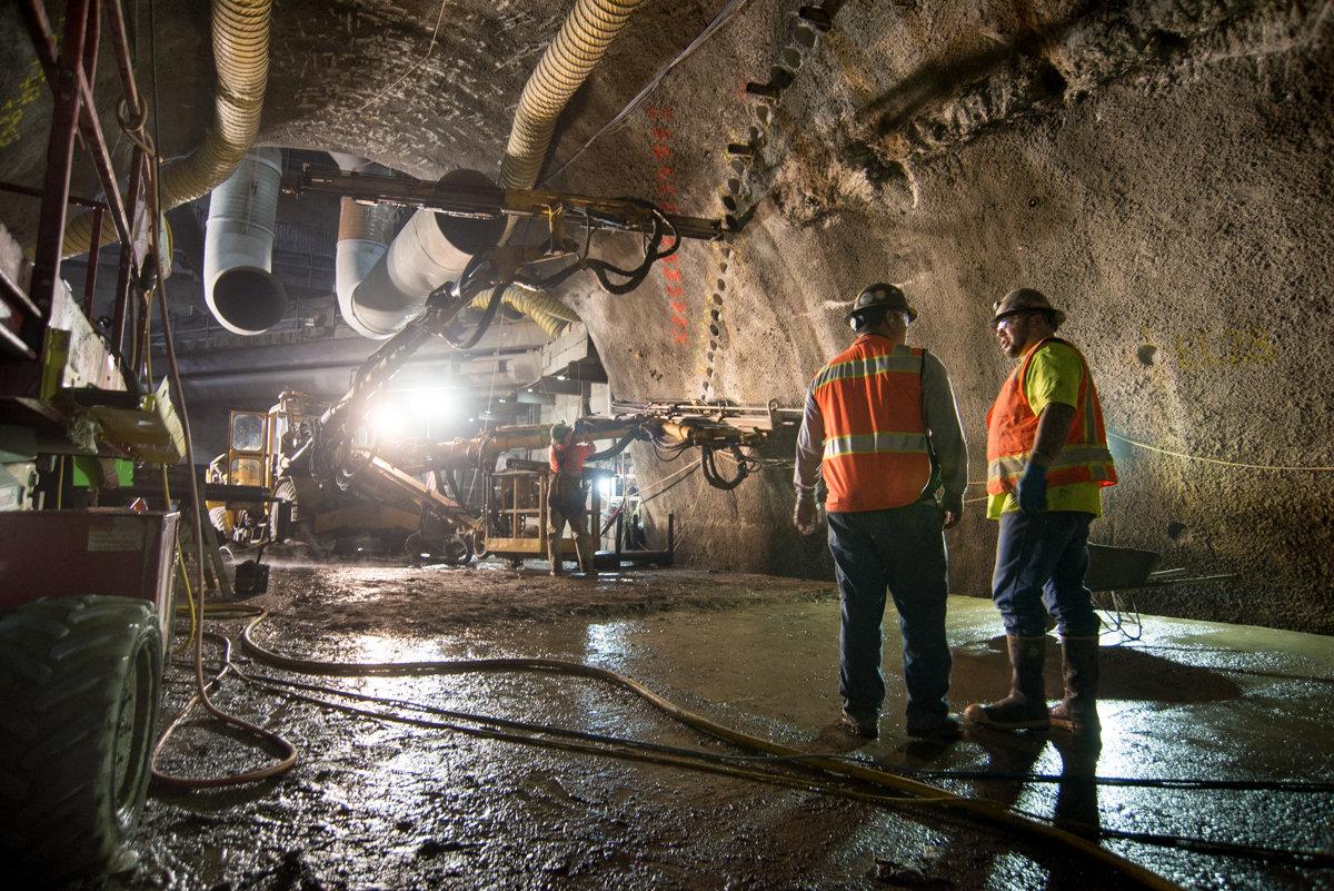 Workers in safety vests observe drilling equipment in underground tunnel during Chinatown Station construction.
