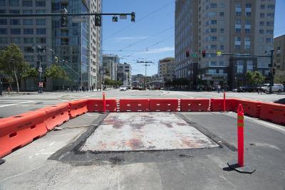 Red traffic barriers block an urban intersection with construction work visible on the street pavement.