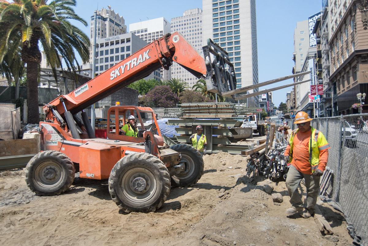 Orange Skytrak forklift lifting equipment at urban construction site on Stockton Street with workers and city buildings in background.