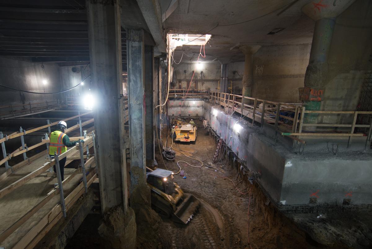 Underground construction site with worker in safety vest, yellow drilling equipment, metal railings, and structural work in progress.