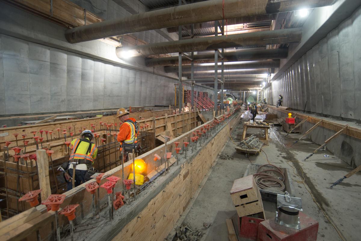 Construction workers in safety gear work on concrete formwork inside an underground transit tunnel.