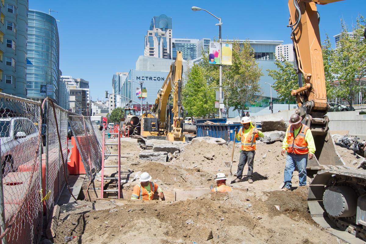 Construction workers and excavators at Yerba Buena Moscone station site during utility relocation and trenching operations.