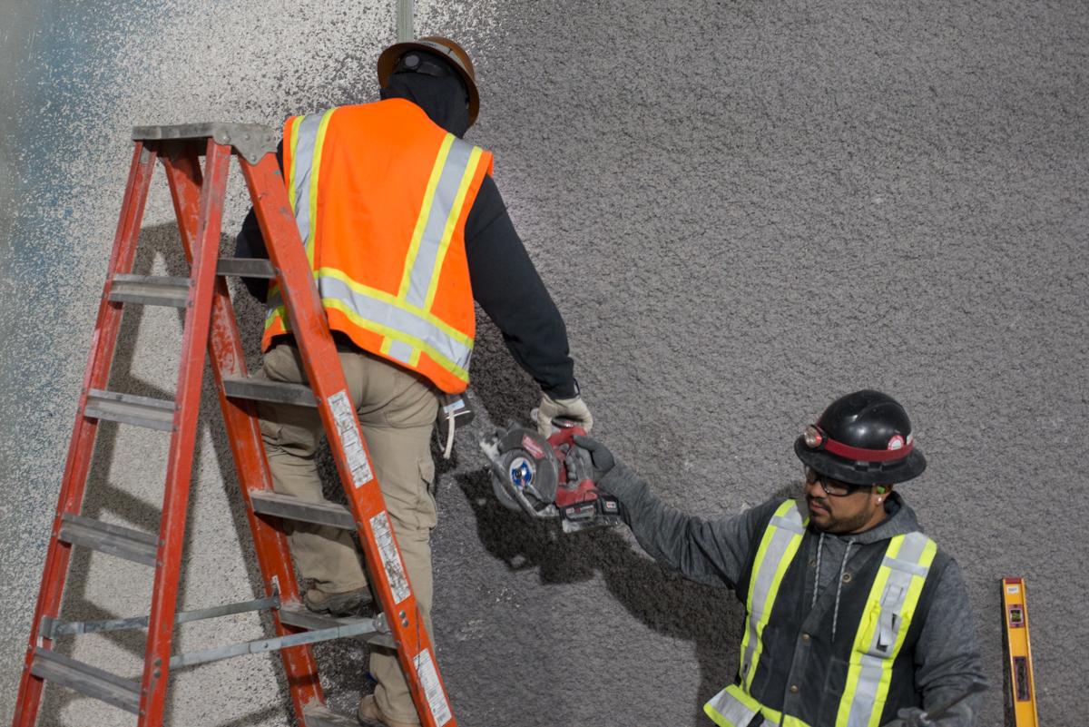 Two workers in safety vests install utilities on a concrete wall using a red ladder and power tools.