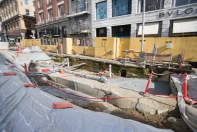 Construction site with yellow equipment, exposed concrete foundation, and red safety barriers in urban area.