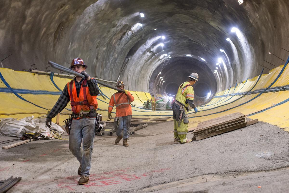 Workers in safety gear inside a tunnel during construction, with yellow protective sheeting and cables along the walls.