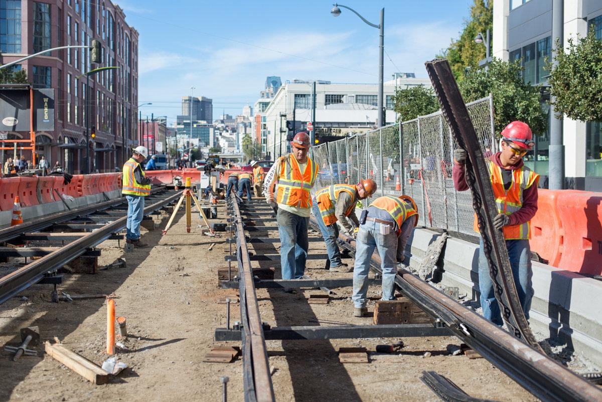 Construction workers in safety gear install railroad tracks on 4th Street with city skyline in background.