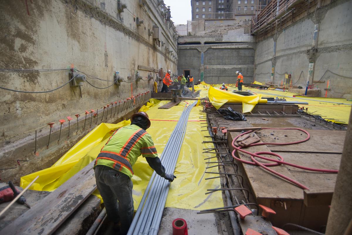 Workers install yellow waterproofing membrane in underground transit construction pit at Union Square.