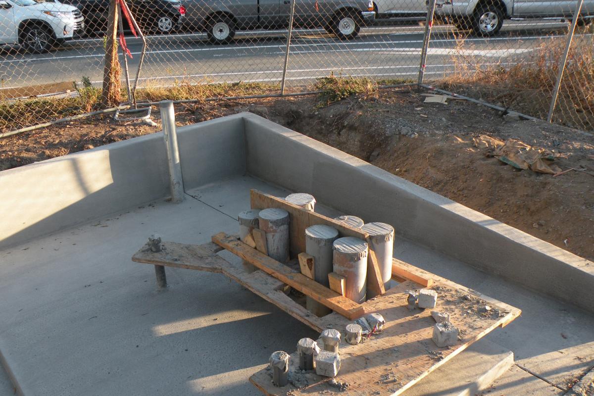 Concrete pit with wooden beam supporting metal conduits and cylindrical components during utility infrastructure installation.