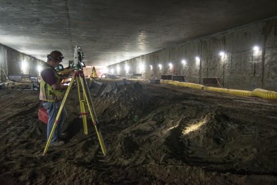Surveyor with tripod measuring underground transit station construction site with concrete walls and lighting.