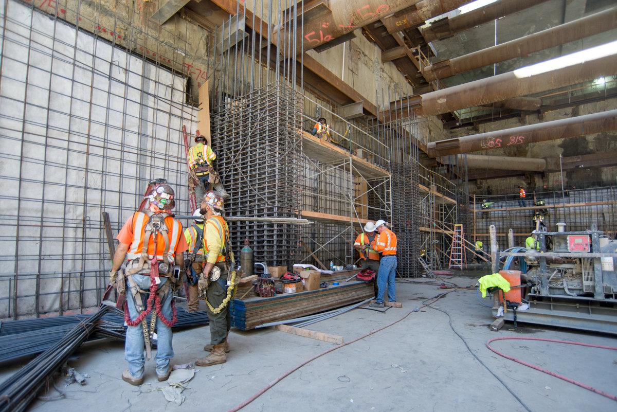 Construction workers in safety gear install rebar cages and waterproofing membranes inside Yerba Buena Moscone Station headhouse.