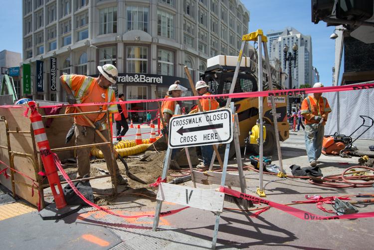 Construction crew works on street restoration at urban intersection with crosswalk closed sign visible.