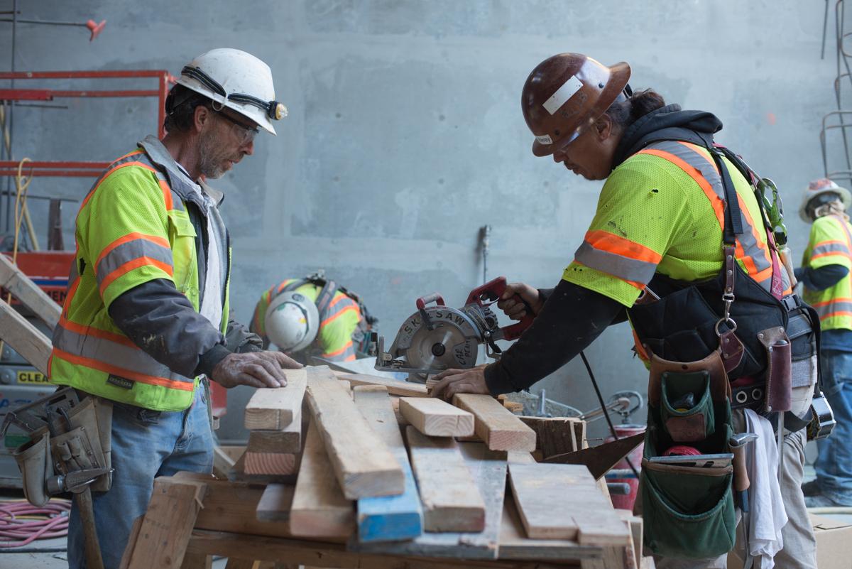 Construction workers in safety gear cut and prepare wooden boards for concrete formwork inside a transit station headhouse.
