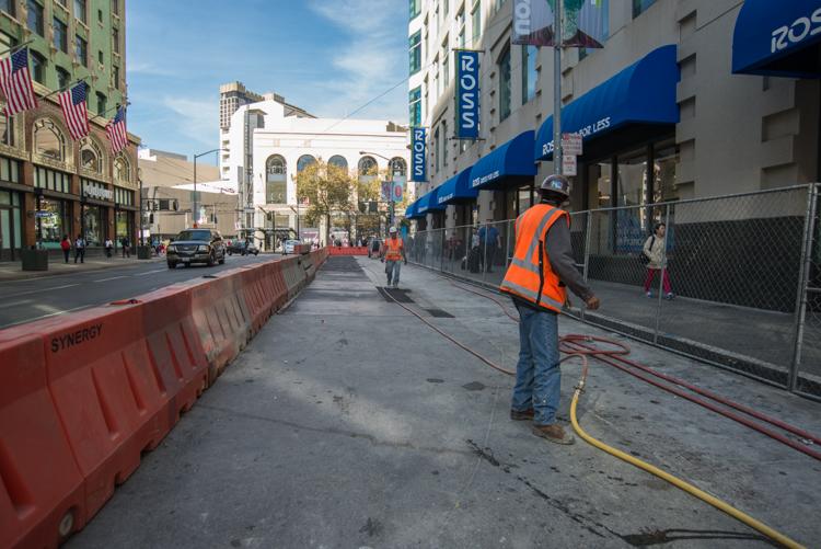 Construction worker in orange safety vest operating equipment on 4th Street with red barriers and blue storefront hoarding visible.